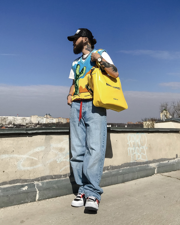Man wearing desert cactus knit sweater vest with jeans and carrying a yellow tote bag outdoors under a blue sky.