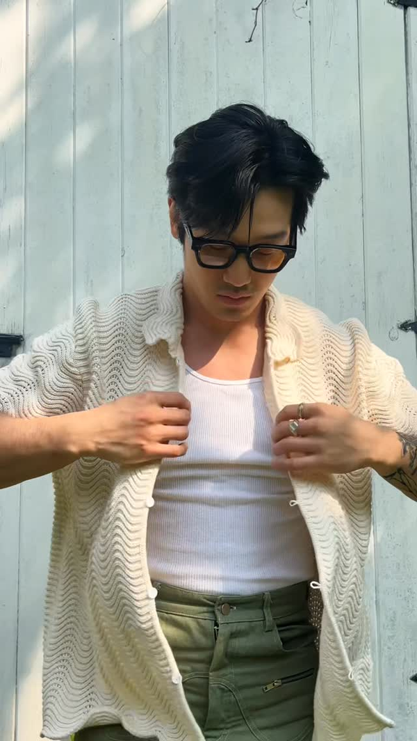 Person wearing a cream shale stitch knit shirt over a white shirt with green pants, standing against a light blue wooden wall.