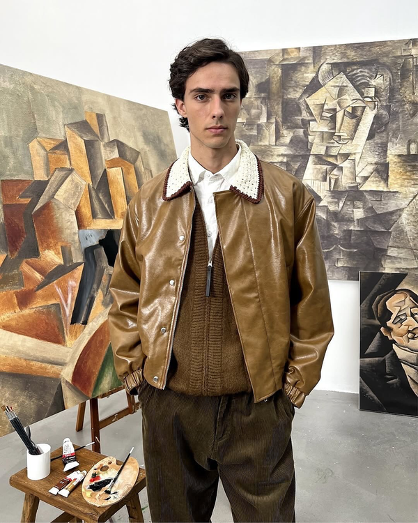 Model wearing a brown leather jacket with crochet collar layered over a knit zip sweater and white shirt in an art studio setting.
